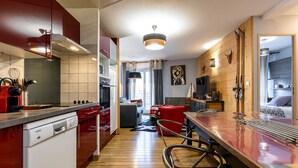A modern kitchen with red cabinetry, a wooden dining area, and a cozy living space with a fireplace. - COZY APARTMENT RENOVATED CITY CENTER AVENUE LECLERC WIFI AND PRIVATE COVERED PARKING (CAUTERETTS)