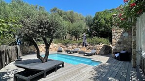 Una terraza de madera con piscina, sillas de descanso y un muro de piedra.