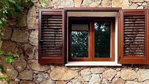 Una casa de piedra con ventana y contraventanas, rodeada de grava y plantas.