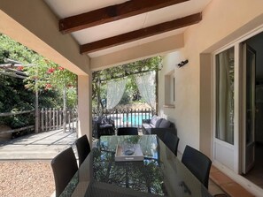 Ein Esstisch mit Glasplatte und Stühlen, Blick auf einen Pool und eine Außenlounge, sowie eine Holzpergola mit roten Blüten.