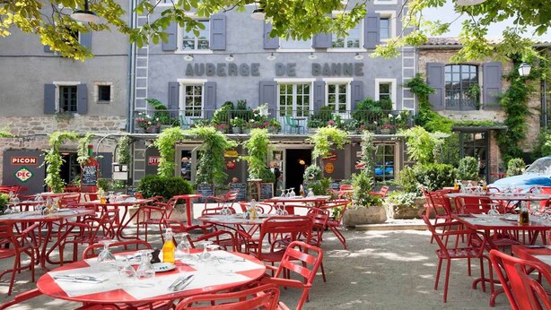 Un área de comedor al aire libre con sillas y mesas de metal rojo, rodeada de follaje y un edificio con la señalización 'AUBERGE DE BANNÉ'.