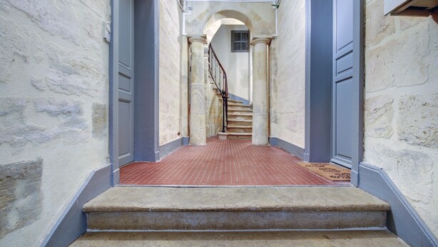 A narrow hallway with a staircase leading to a landing. The walls are textured and painted in a light color, with a red-tiled floor.