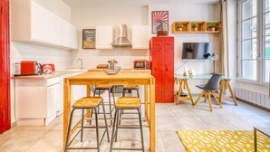 A modern kitchen with white cabinets, a wooden dining table with stools, a red cabinet, and a TV on a shelf.