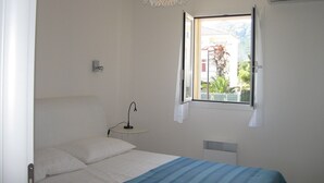 A bedroom with a bed, a bedside table with a lamp, a window with a view of buildings and greenery, and a ceiling light fixture.