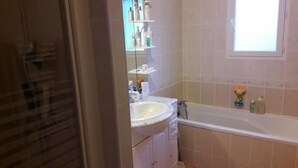 Un baño con bañera, lavamanos y espejo. Hay artículos de aseo en los estantes.
