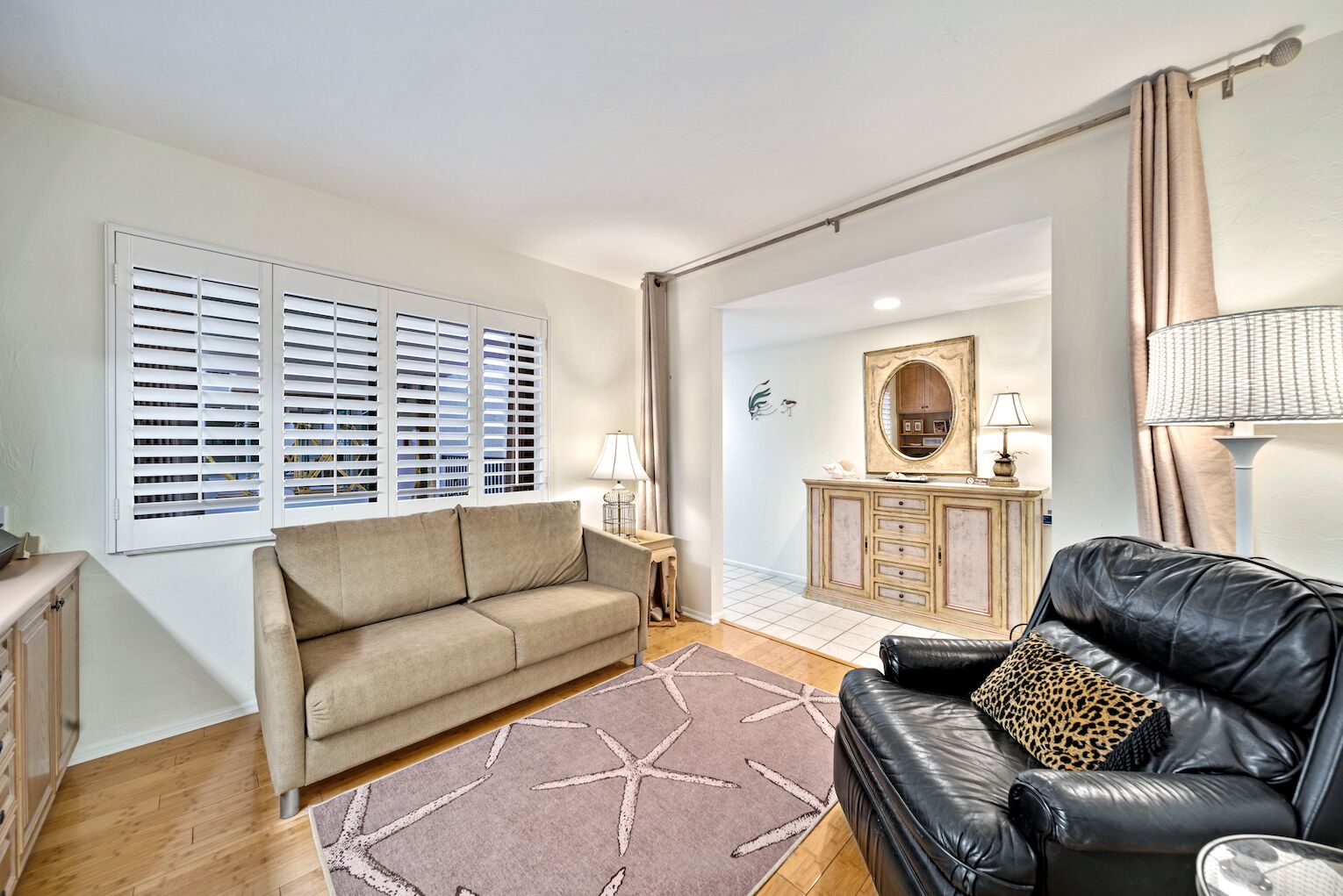Un salon avec un canapé beige, un canapé en cuir noir, un tapis orné de motifs d’étoiles de mer, un meuble en bois et un miroir au mur.