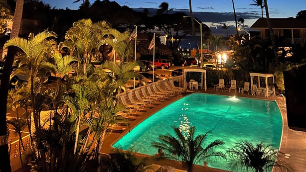 A nighttime view of a pool area with lounge chairs and palm trees. - KIHEI KAI NANI #6-246  Beautiful open view with  Ocean/Pool in Tropical Garden (Kihei)