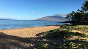 A beach with a person walking, clear blue sky, and a mountain in the background. - KIHEI KAI NANI #6-246  Beautiful open view with  Ocean/Pool in Tropical Garden (Kihei)