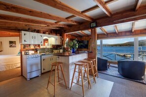 Eine rustikale Küche mit Holzbalken, einem Herd, Barhocker und Seeblick durch große Fenster.