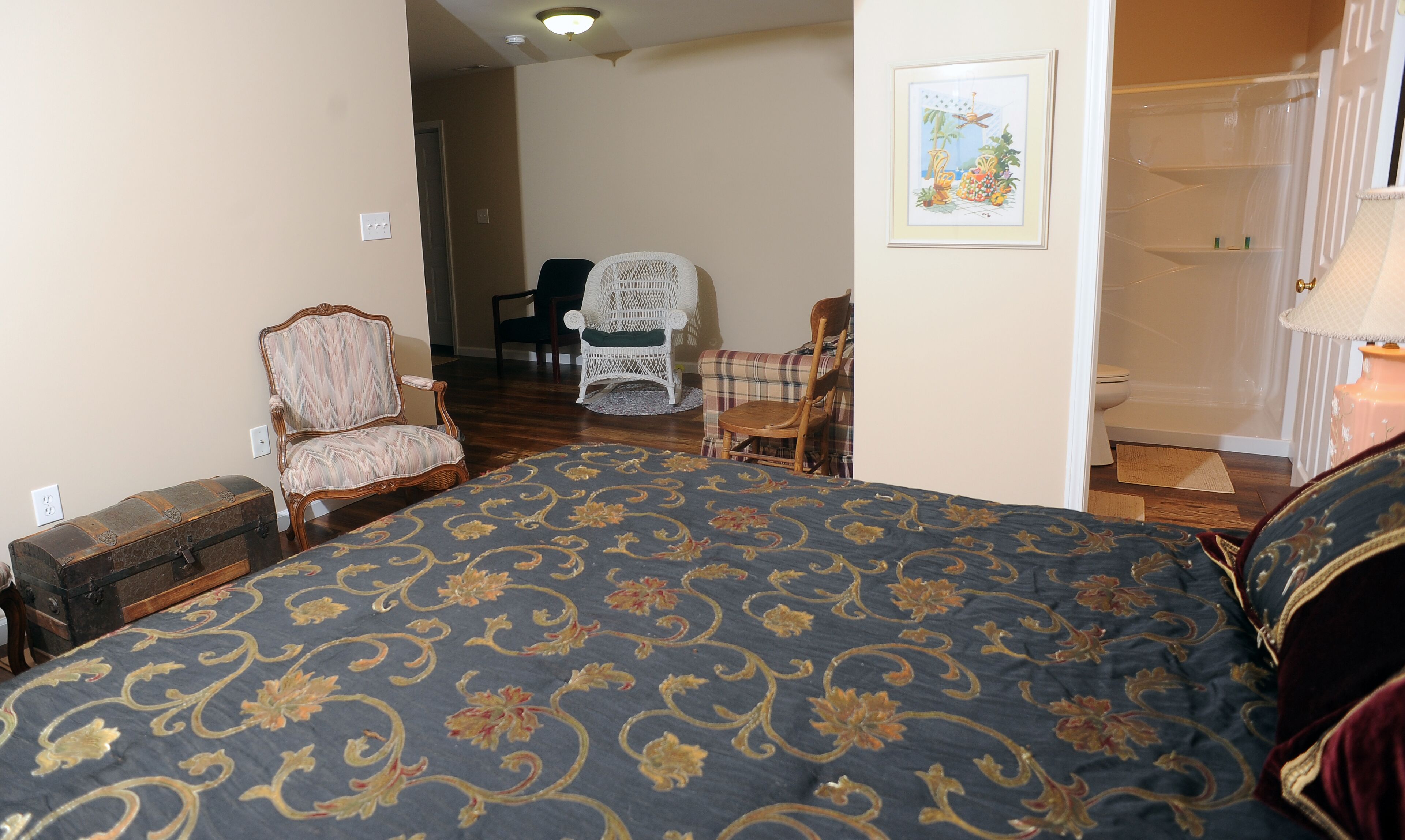 Un dormitorio con una cama grande, un cofre de madera, una silla de mimbre, un sillón de cuadros y un cuadro en la pared.