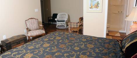 Un dormitorio con una cama grande, un cofre de madera, una silla de mimbre, un sillón de cuadros y un cuadro en la pared.