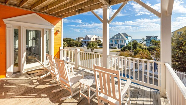 A wooden deck with white rocking chairs, a glass door entrance, and a view of neighboring houses. - Dreamsicle: Pool, Pup & Family Fun (Emerald Isle)