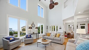 A spacious living room with large windows, a sofa set, a coffee table, and a dining area. - Dreamsicle: Pool, Pup & Family Fun (Emerald Isle)