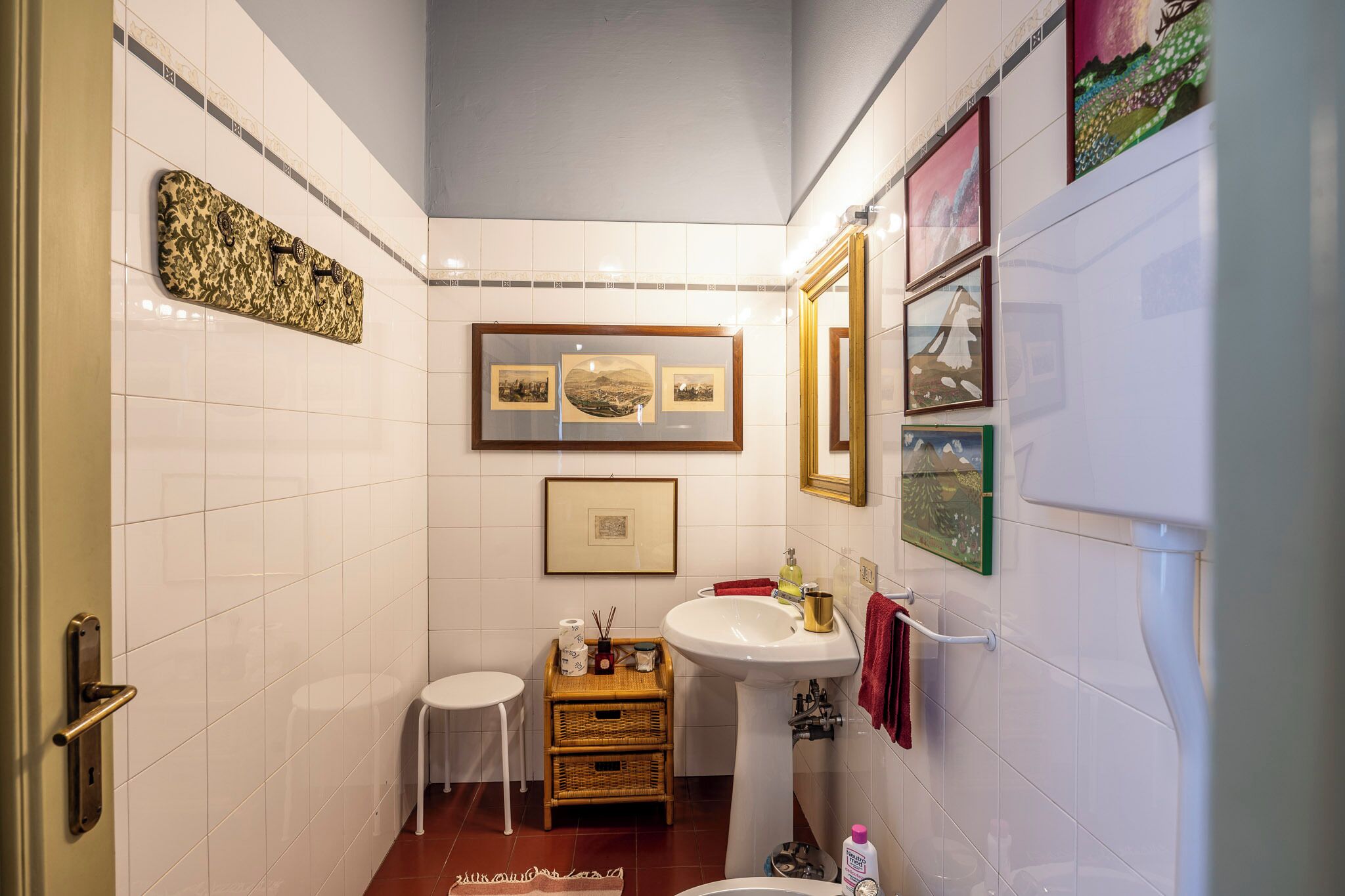 A bathroom with white tiled walls, a white sink, a small stool, and framed pictures on the wall.