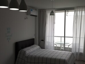 A bedroom with a bed, a wall-mounted air conditioner, a ceiling light, a window with sheer curtains, and a small table by the window. - Independencia 1302 9C by HB (Buenos Aires)