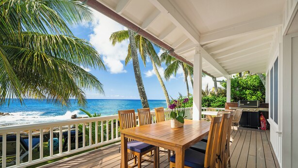 Une terrasse en bois avec une table et des chaises, offrant une vue sur l’océan et bordée de palmiers.
