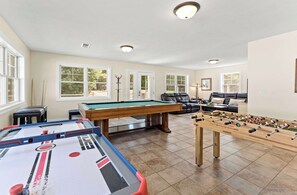 A pool table, foosball table, and a billiards table in a well-lit room with large windows.