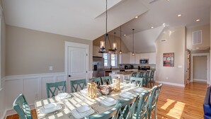 A spacious dining area with a large table set for six, surrounded by teal chairs. A chandelier hangs above, and the room has a high ceiling with recessed lighting. - 5547- Sailfish Station - Nags Head Luxury Home Private Pool OBX (Nags Head)