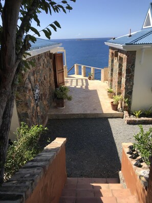 A coastal property with a stone wall, a wooden door, and a paved pathway leading to a balcony overlooking the sea.