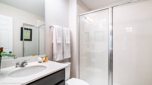 Une salle de bain moderne avec une douche à parois de verre, un grand miroir et un lavabo blanc agrémenté d’articles de toilette.