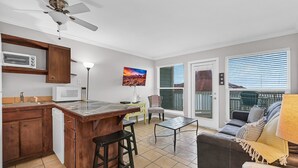 A kitchen with a microwave, wooden cabinets, and a bar stool. There is a dining area with a table and chairs, and a sofa with a lamp.