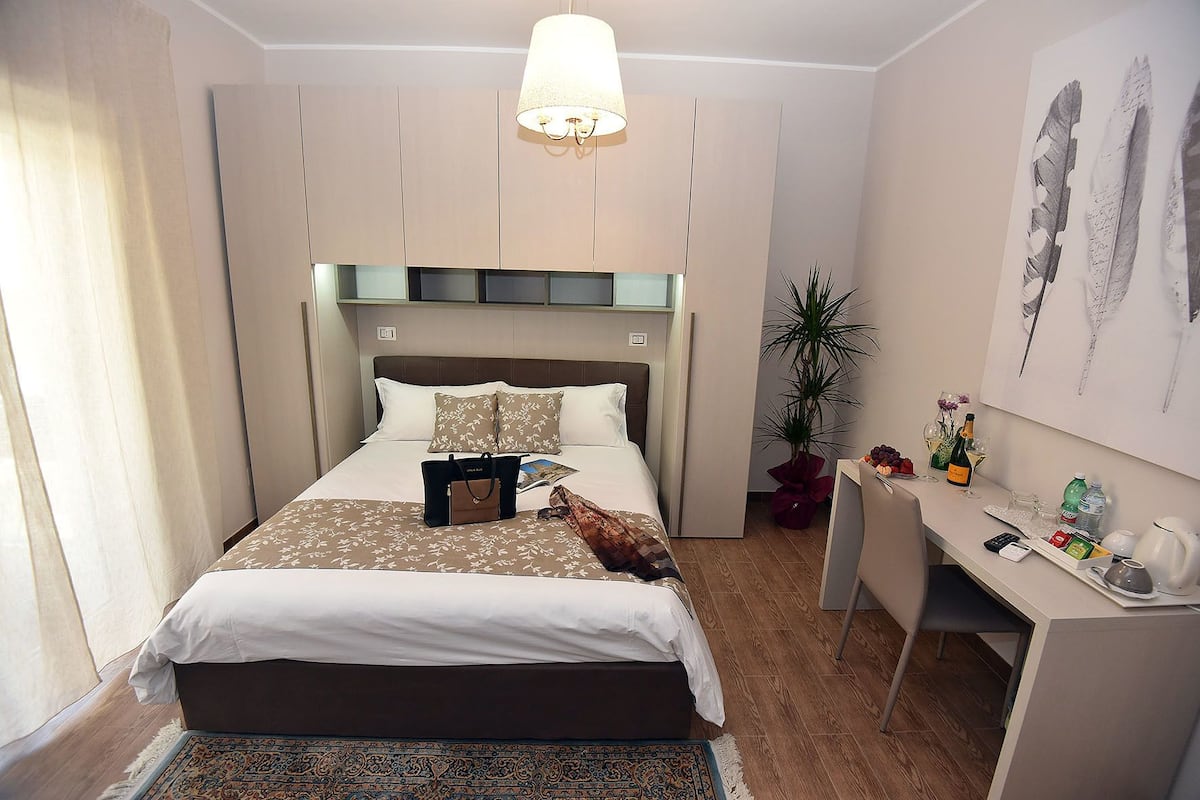A hotel room with a bed, a desk, a lamp, a plant, and artwork on the wall.