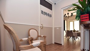 Un couloir avec une chaise en bois, un panneau fixé au mur et un coin repas visible à travers une porte ouverte.