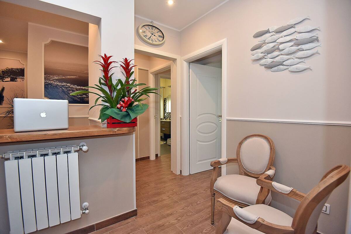 A reception area with a wooden reception desk, a laptop, a potted plant, a clock, and a decorative wall piece.