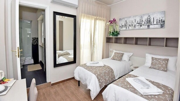 Une chambre d’hôtel avec deux lits, un grand miroir, un tableau représentant un paysage urbain et une salle de bain visible à travers une porte ouverte.