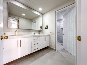 A modern bathroom with a large mirror, white cabinets, and a shower area visible through an open door.