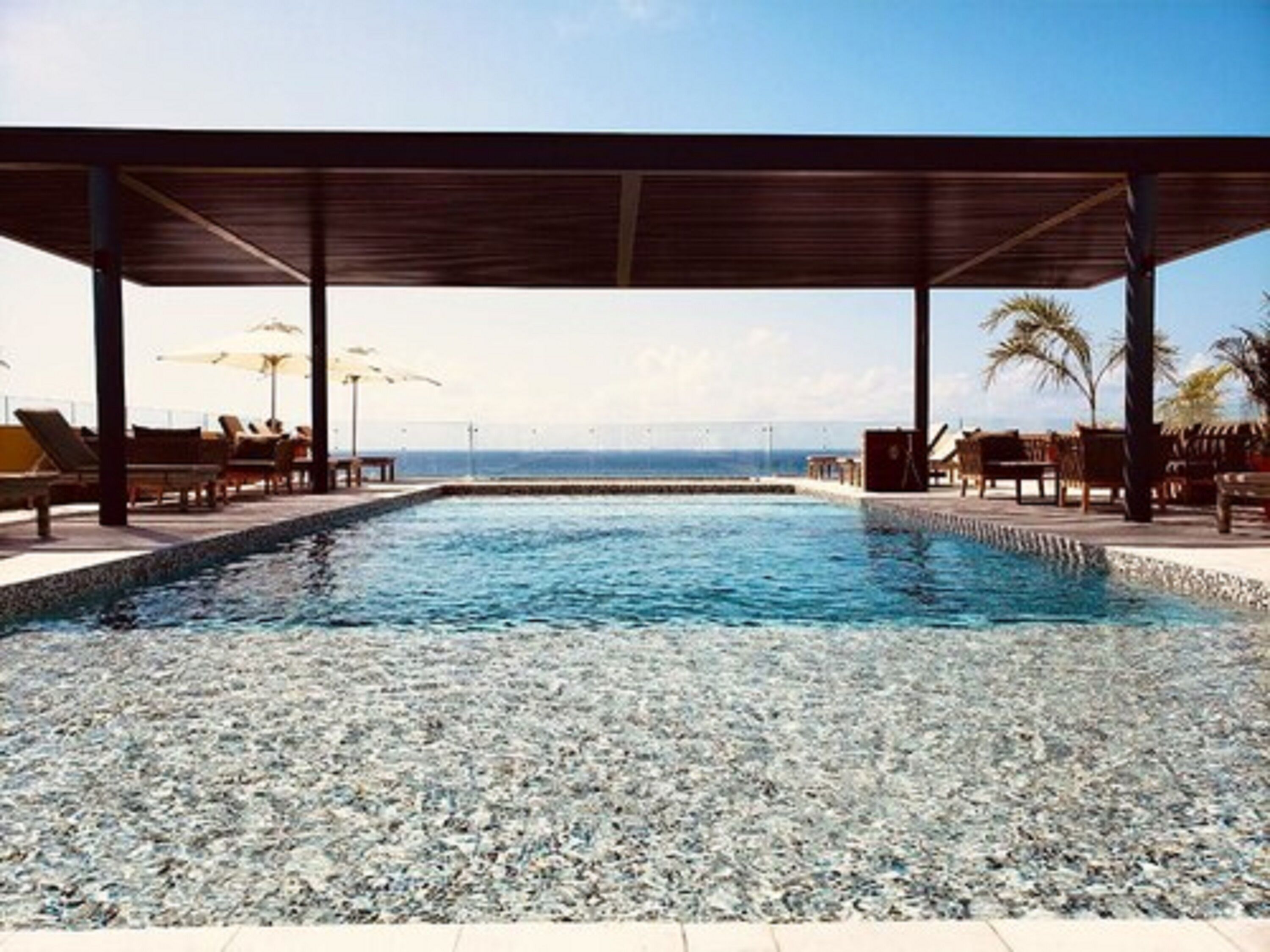 A pool area with lounge chairs and umbrellas, a covered seating area, and a view of the ocean.