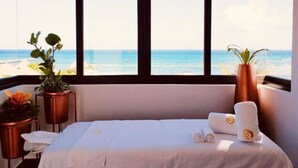 A hotel room with a large window offering a view of the beach and ocean. There is a bed with white linens and a rolled towel on it. Two potted plants are placed on either side of the bed. - Singular Joy Downtown Residences (Playa del Carmen)