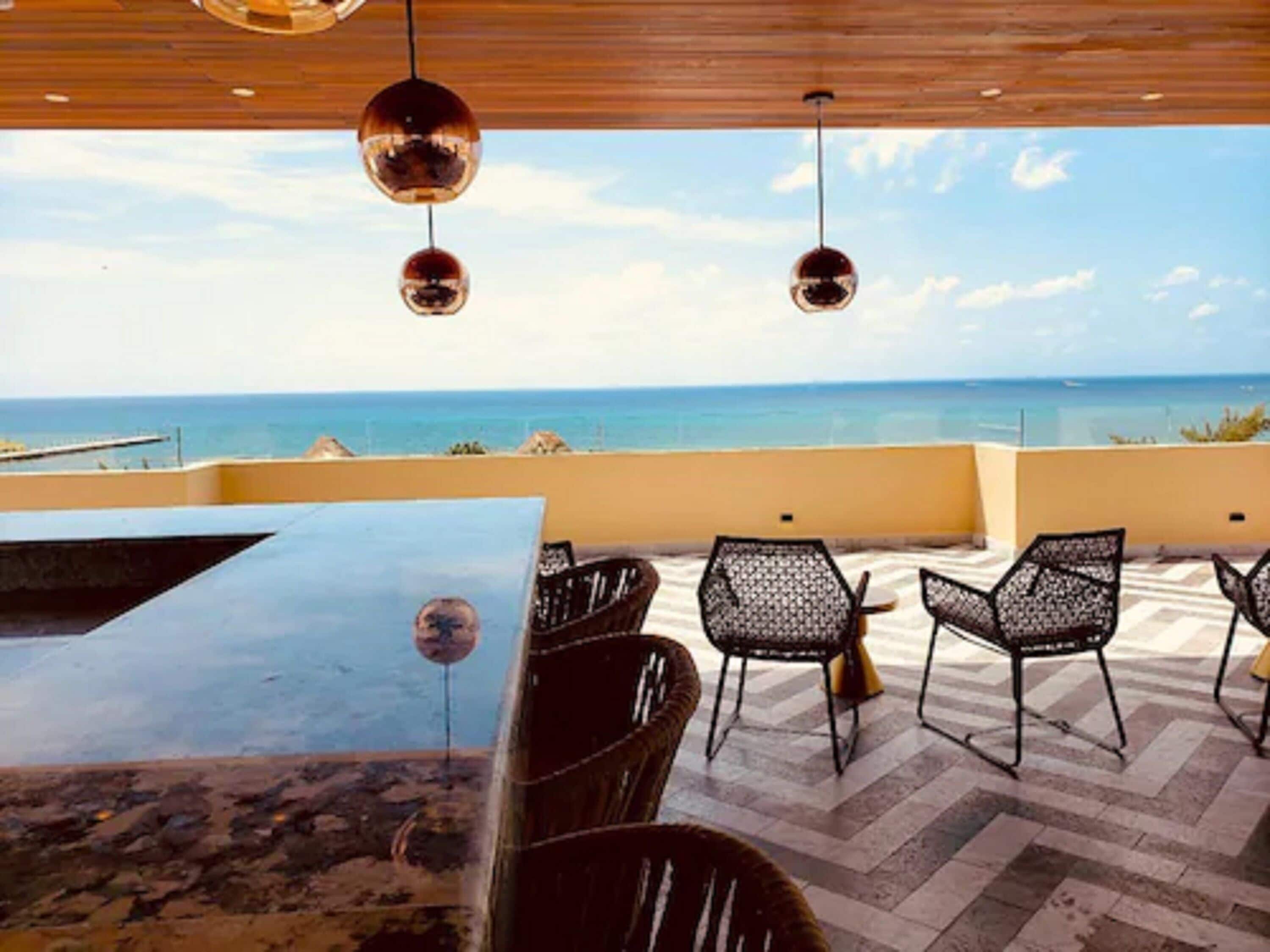 An outdoor seating area with a view of the ocean, featuring a wooden ceiling, hanging spherical lights, and patterned chairs.