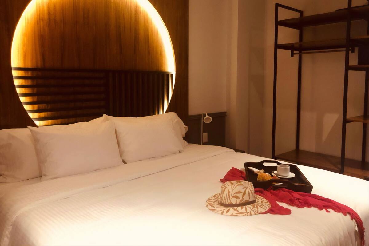 A neatly made bed with white linens, a circular wall light, a small bedside table with a cup, and a red throw blanket.