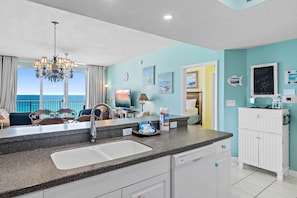 A kitchen with a granite countertop, white cabinets, a stainless steel sink, and a view of the ocean through large windows. - Beautiful Gulf Views⛱️-Indoor/Outdoor Pools- Lucky 7🔆 (Miramar Beach)
