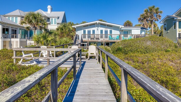 Une promenade en bois mène à une propriété située en bord de mer, comprenant plusieurs maisons, une terrasse couverte et un espace salon extérieur.