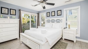Une chambre à coucher avec un lit, des tables de chevet blanches, un ventilateur de plafond et une fenêtre avec des stores.