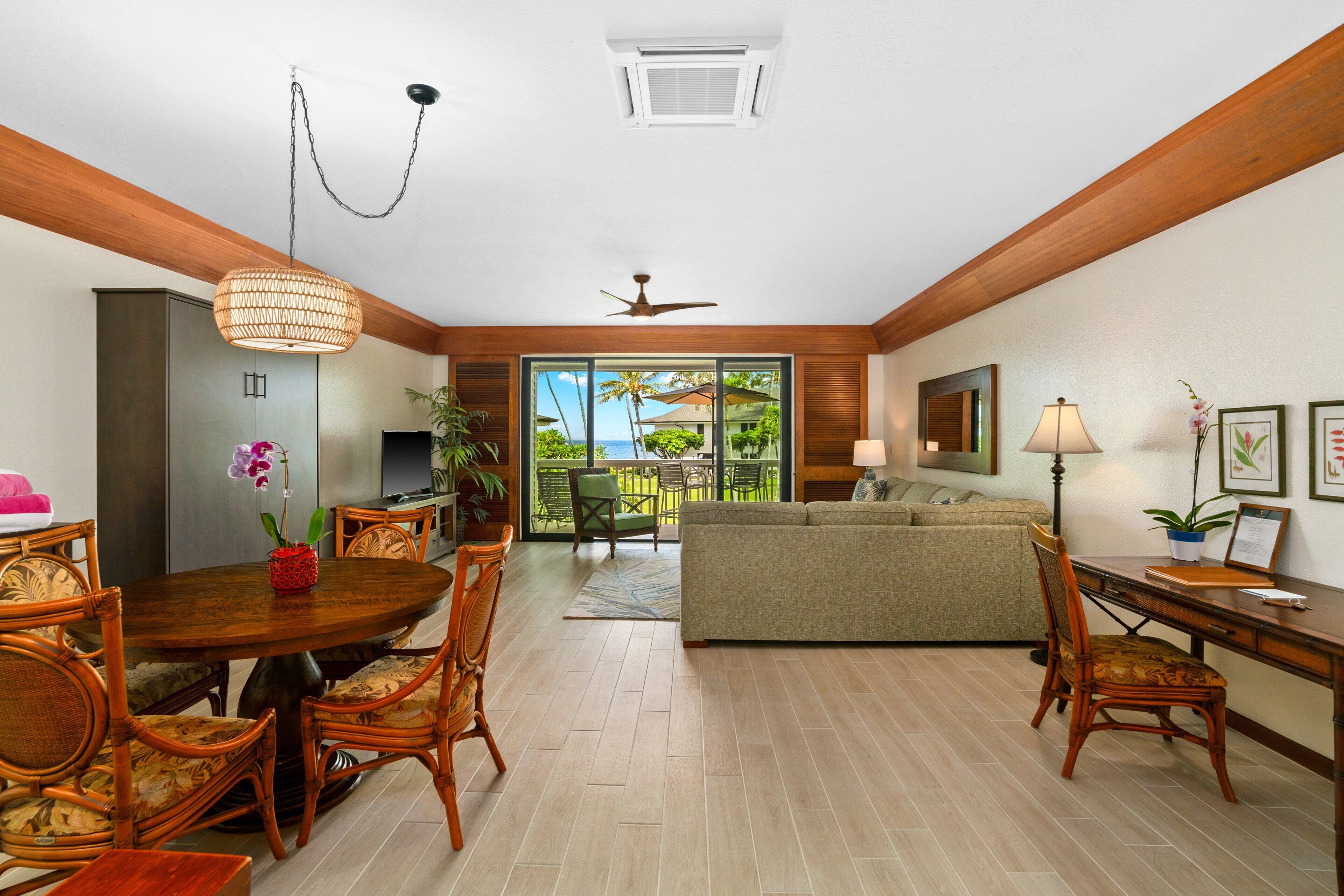 A spacious living area with a dining table, chairs, a sofa, and a desk. There is a ceiling fan, a hanging light fixture, and a view of the outdoors through large windows.