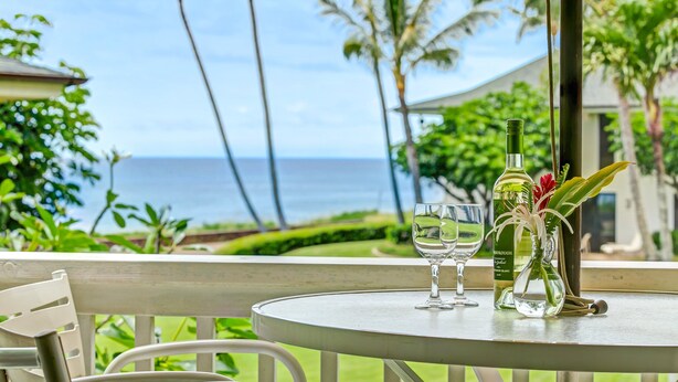 Ein Tisch mit einer Weinflasche, einem Glas und einer Blumenvase, vor einer Aussicht auf das Meer und Palmen.