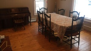 A dining room with a wooden table, six chairs, a piano, and two windows with curtains.