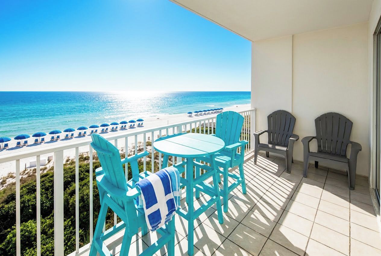 A balcony with a view of the beach, a table, chairs, and umbrellas.