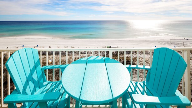 Vista a la playa con sillas turquesa y una mesa en una terraza de madera.