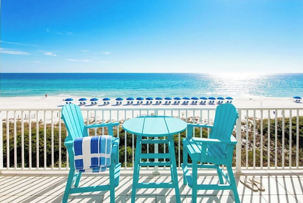 A balcony with turquoise chairs and a table, overlooking a beach with umbrellas.