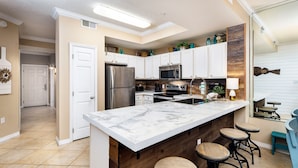 Una cocina moderna con armarios blancos, encimera de mármol, refrigerador de acero inoxidable, microondas y lavabo. Hay tres taburetes de madera en la zona del mostrador.