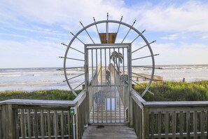 Une promenade en bois menant à une plage, avec une barrière métallique ornée d’un motif représentant un palmier.
