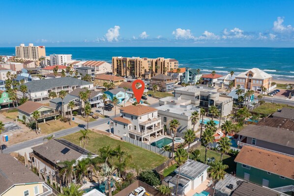 An aerial view of a coastal residential area with multiple buildings, including a prominent hotel with a pool and a view of the ocean.