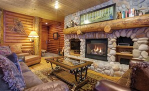 L’intérieur d’un chalet confortable, avec des murs en rondins de bois, une cheminée en pierre, des meubles en cuir et un téléviseur fixé au mur.