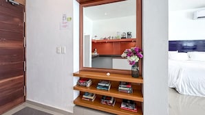 A hotel room with a wooden door, a large mirror, a bookshelf with books, a vase with flowers, and a bed with white bedding.