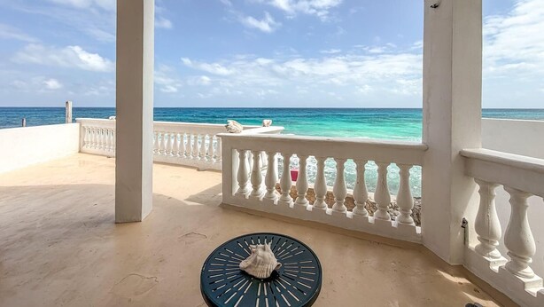 A covered balcony with a view of the ocean, a white railing, and a round table with a shell.