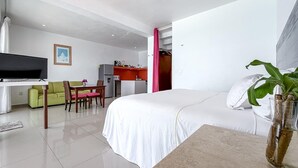A hotel room with a bed, a TV, a dining area with a table and chairs, a kitchen area, and a plant on a table.